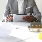Selective focus. Design and architecture. Cropped shot of African architect holding digital tablet, developing new real estate project, sitting at desk with architectural tools and scale model house
