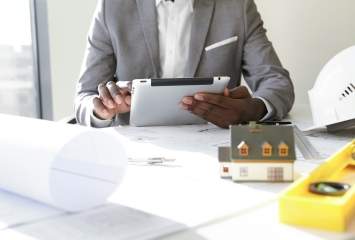 Selective focus. Design and architecture. Cropped shot of African architect holding digital tablet, developing new real estate project, sitting at desk with architectural tools and scale model house